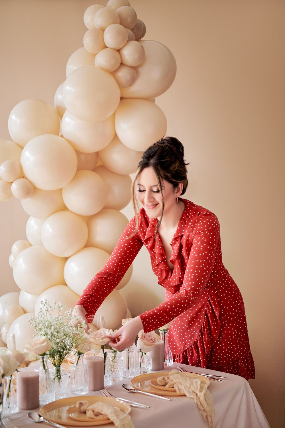 Event planner in red polka dot dress arranges table decor next to elegant white balloon arch display.
