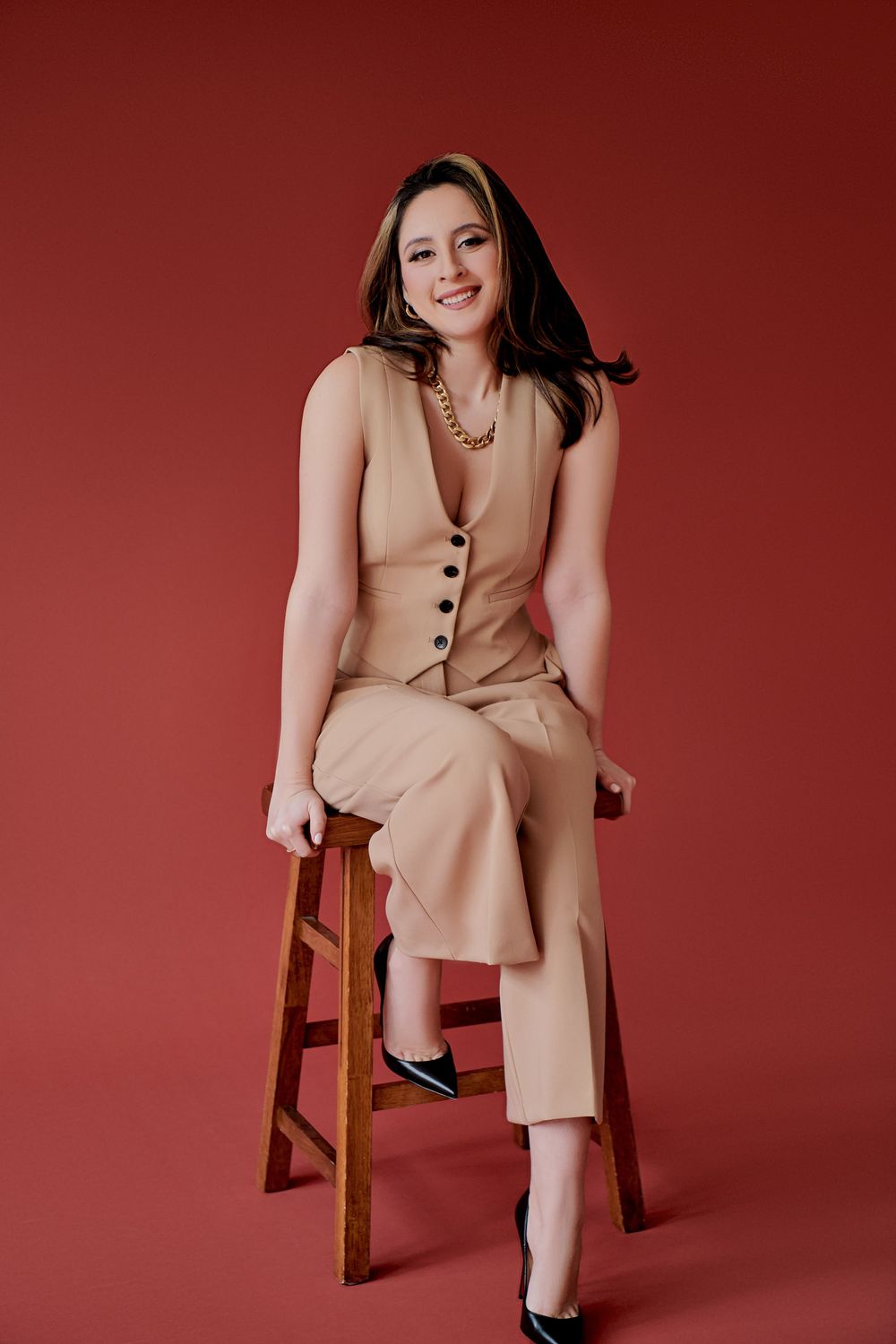 Professional studio portrait of someone wearing a beige pantsuit and heels seated on wooden stool against red background.