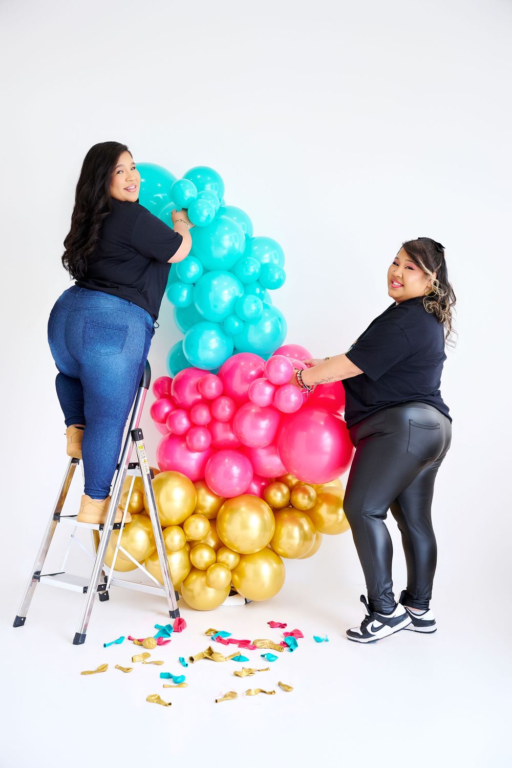 Two women arranging a colorful balloon garland with teal, pink, and gold balloons on a white background.