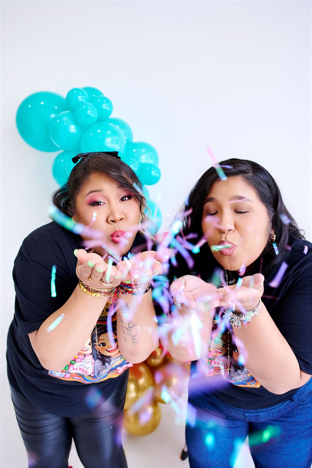 Two women blowing colorful confetti with teal balloons in background at a festive celebration party.