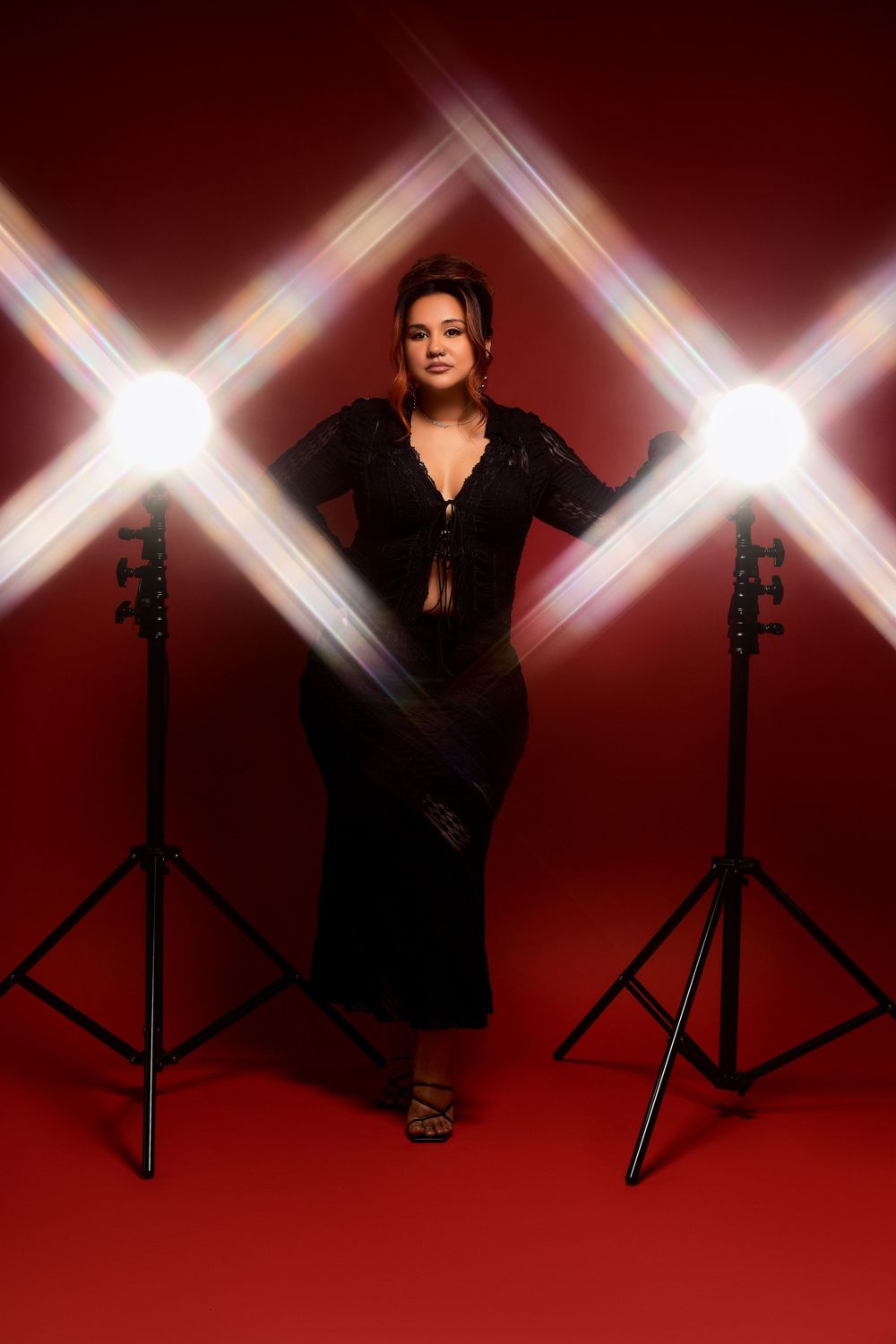 A figure in a black dress poses confidently between two light stands against a dramatic red background with crossed light beams.