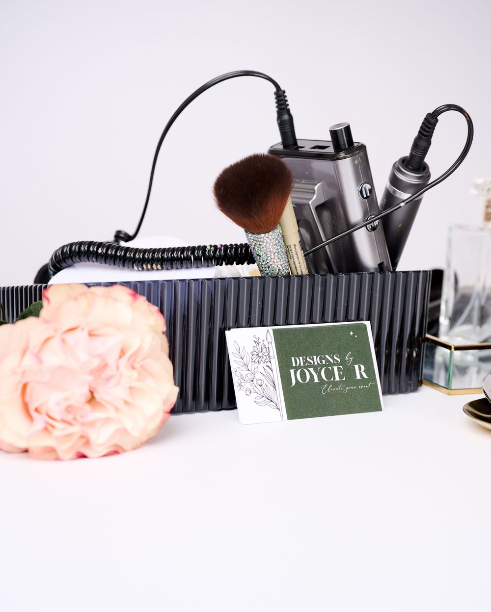 Makeup brush, hair tools, and a Designs by Joyce R business card displayed with a pink peony on a white surface.