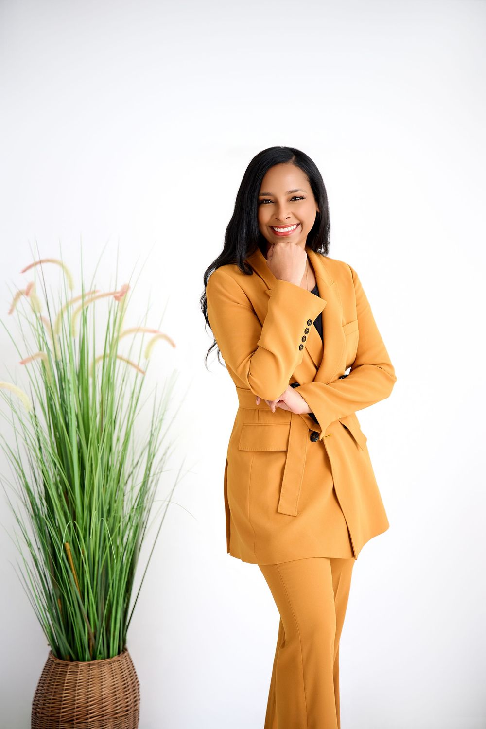 Woman in mustard yellow suit smiling confidently, standing next to a tall potted grass plant on white background.
