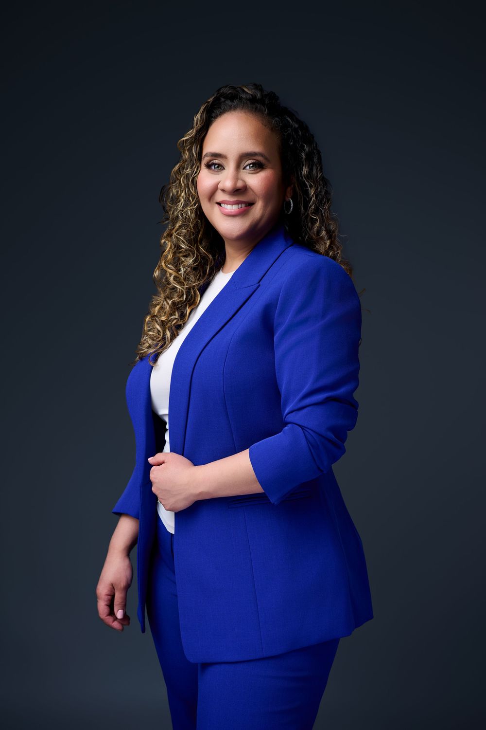 A professional woman in a royal blue suit smiles confidently against a dark gray studio background. Headshot photographer new jersey