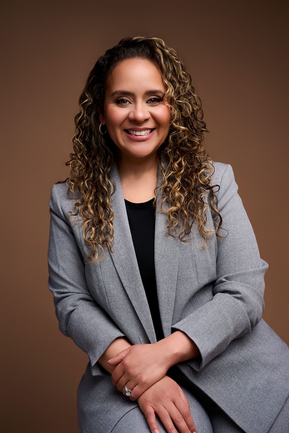 Professional woman in gray blazer with curly highlighted hair smiling confidently against brown background. NJ headshot photographer