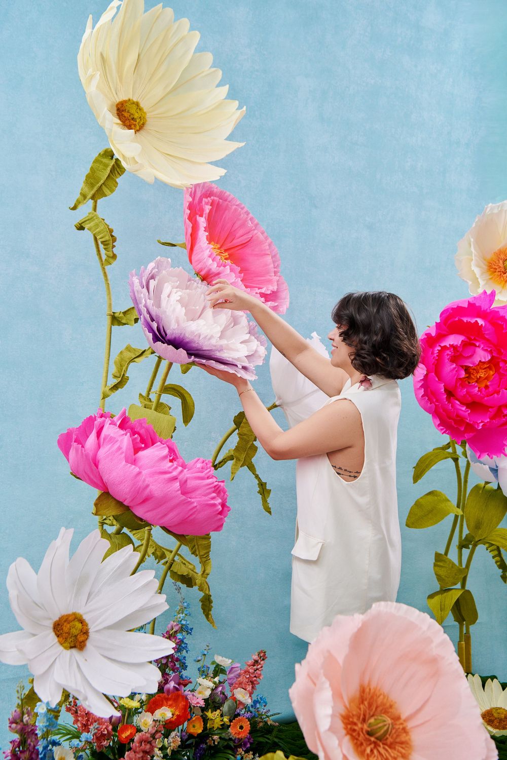 Collage of colorful giant paper flowers against a blue background with someone reaching up to arrange white and pink blooms.