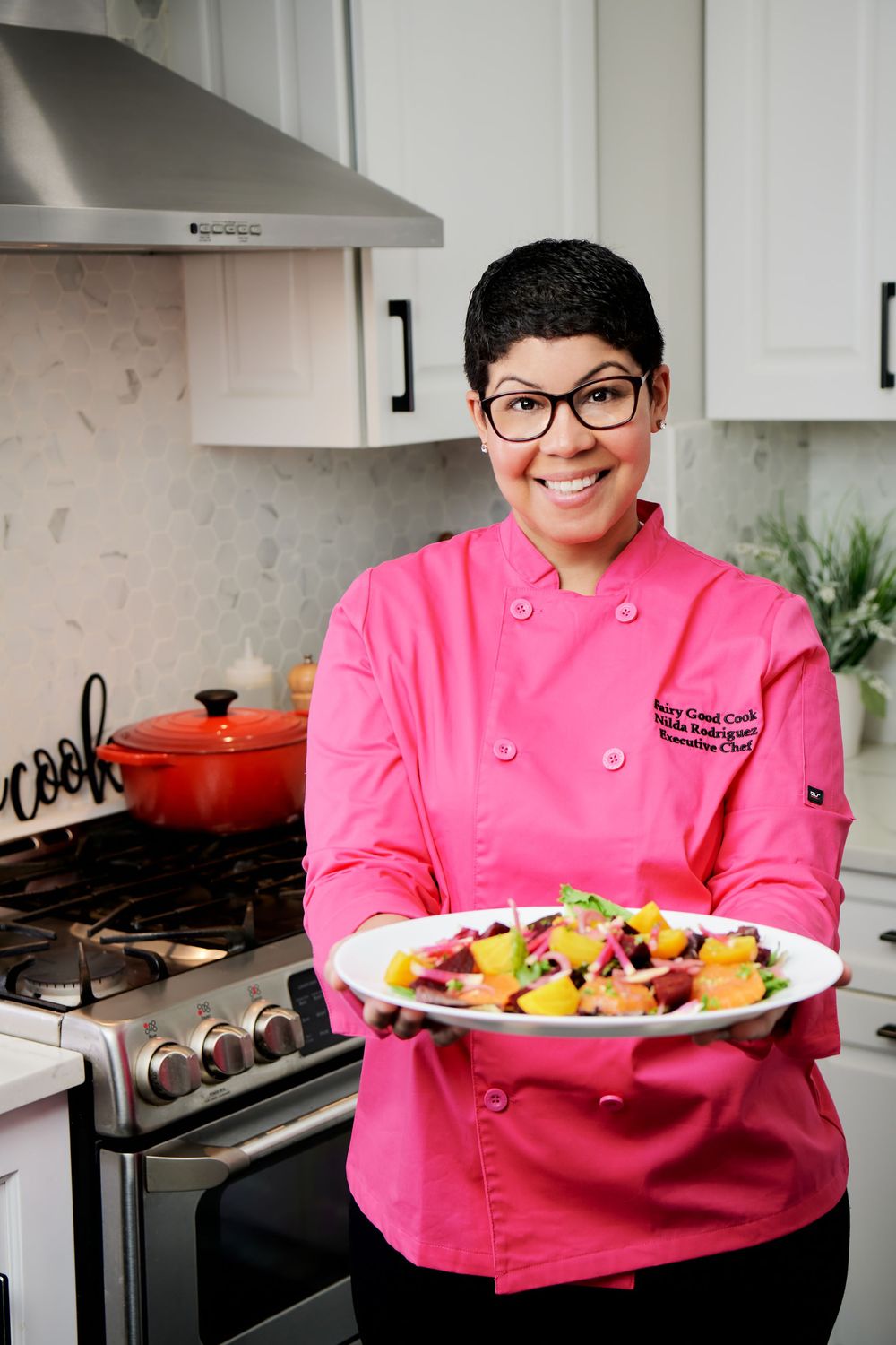 Chef in pink jacket presents colorful plated dishes in modern white kitchen.