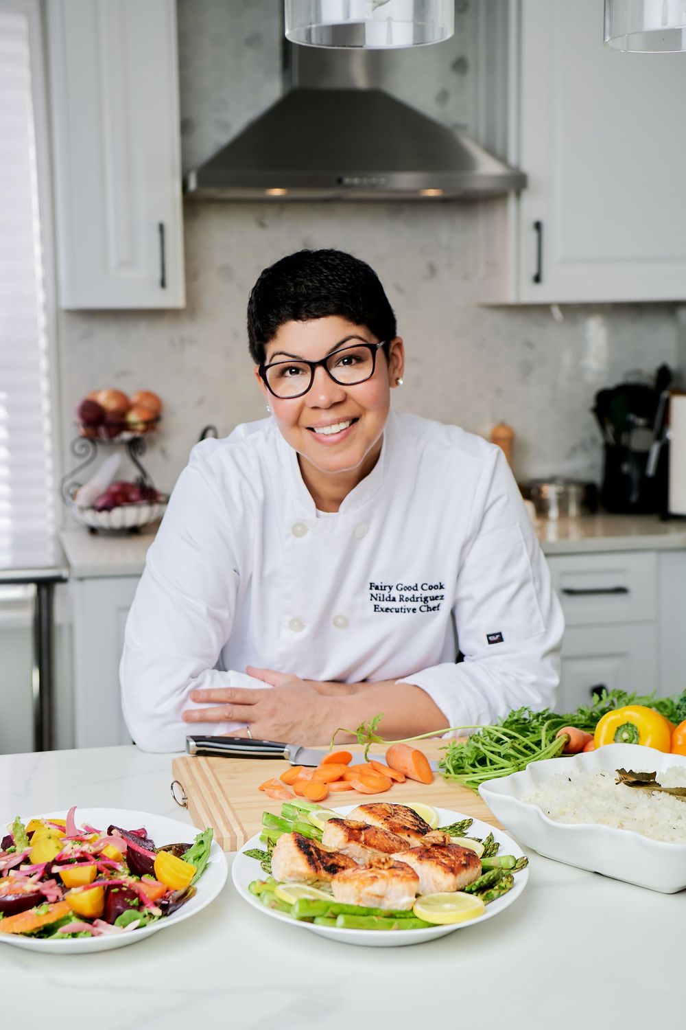 Chef displays variety of fresh ingredients and prepared dishes on white kitchen counter.
