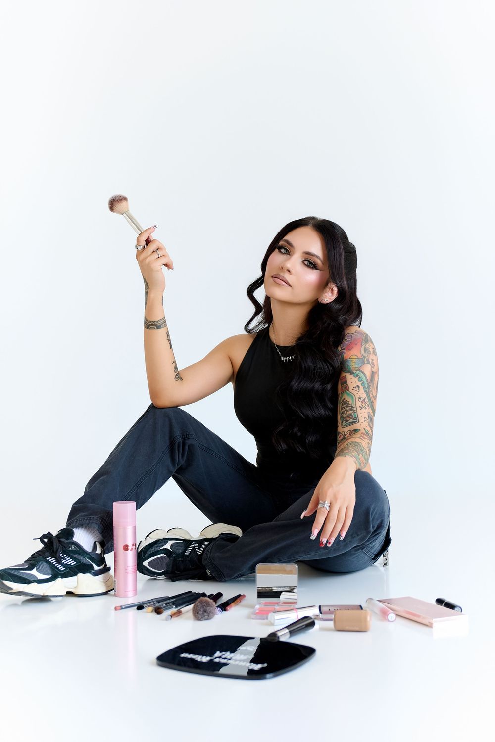 Tattooed woman sitting on white floor holding makeup brush surrounded by cosmetics products wearing black top and jeans.