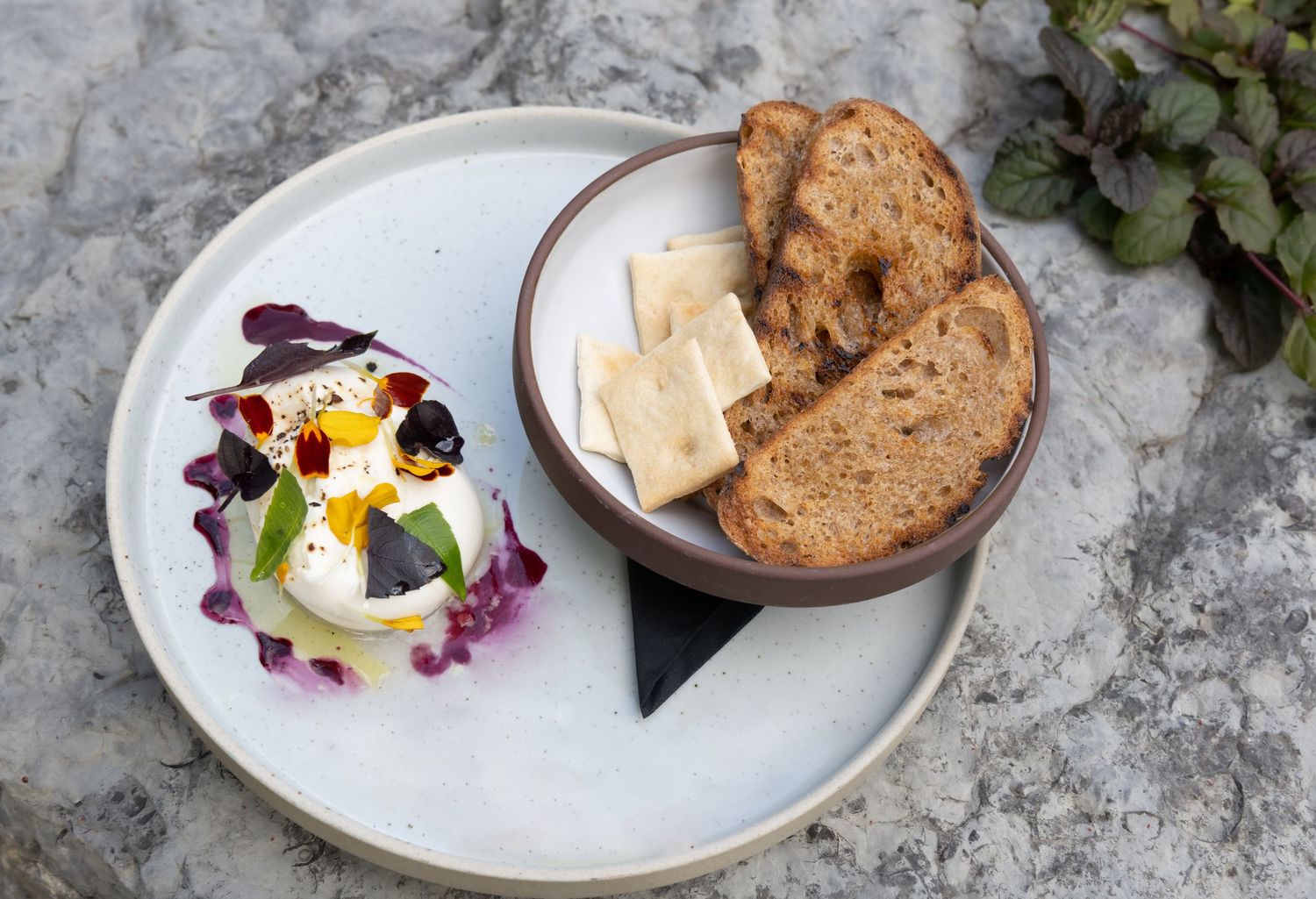 Artfully plated bread service with poached egg and purple edible flowers on grey stone surface. The Manse Picton