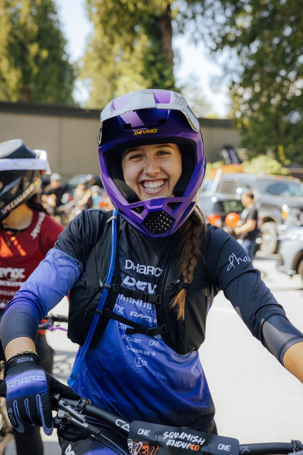Mountain biker wearing purple helmet and blue jersey poses with their bike at an outdoor event.