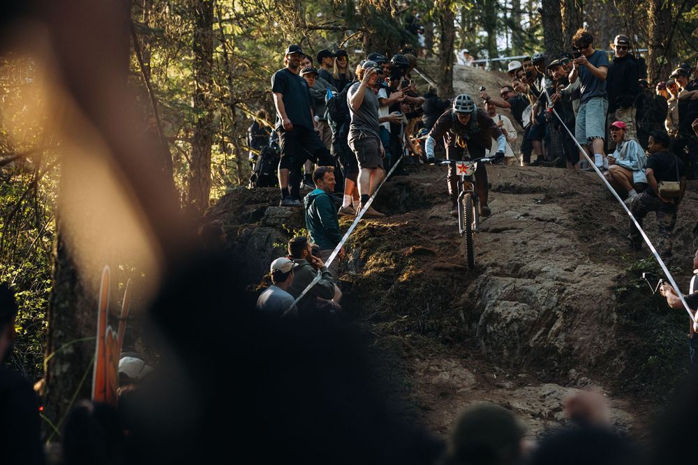 Mountain bike rider navigating rocky terrain on forest trail while spectators gather to watch during competition.