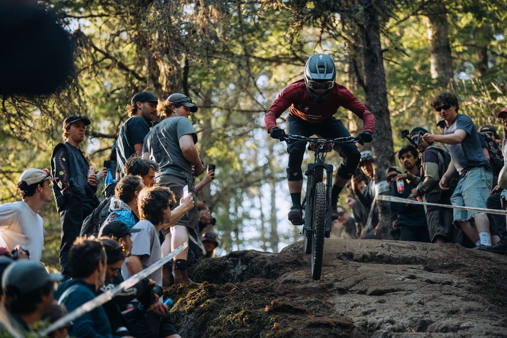 Mountain biker performs aerial jump on dirt trail while spectators watch from sidelines during downhill competition.