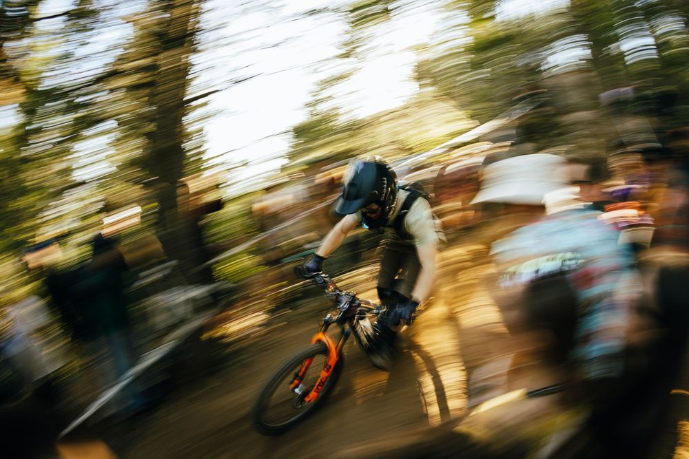 Mountain biker races downhill at high speed during competition with motion blur effect and spectators in background.