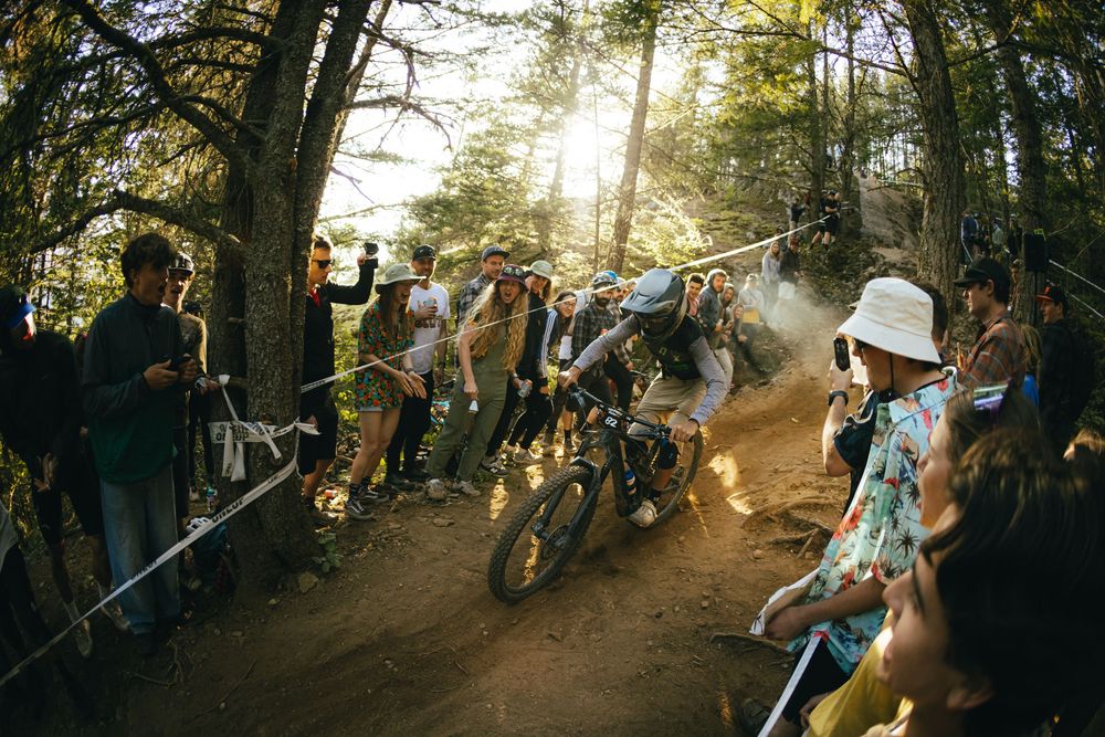 Mountain bikers race through dusty forest trail as spectators watch during downhill competition in sunlit woods.