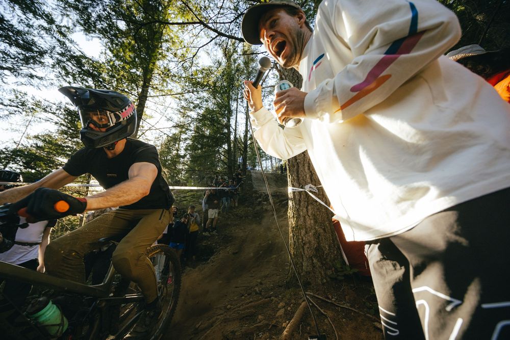 Two mountain bikers film video content while riding on a dirt trail through forest terrain in natural lighting.