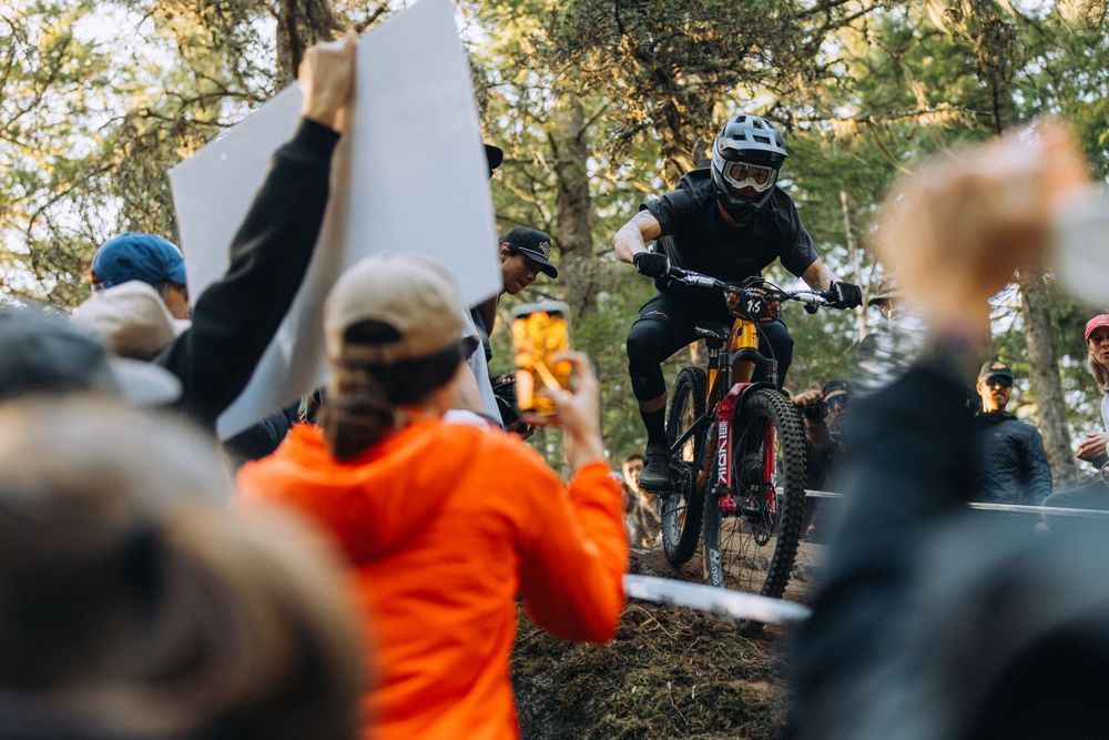 Mountain bikers compete on a forest trail while spectators cheer from the sidelines.