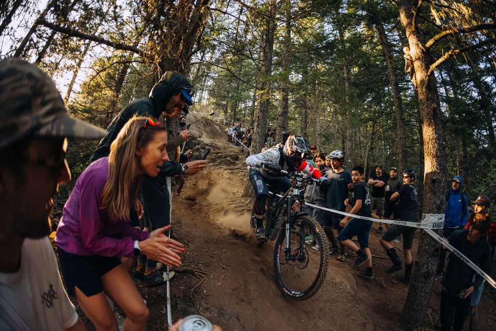 Mountain bikers gather around a trail section to watch and encourage fellow riders attempting technical features.