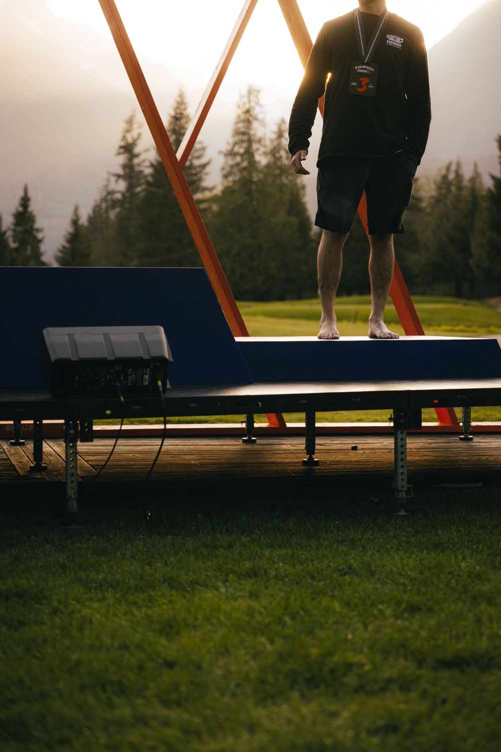 Mountain biker stands silhouetted on a wooden ramp during sunset with evergreen trees in the background.