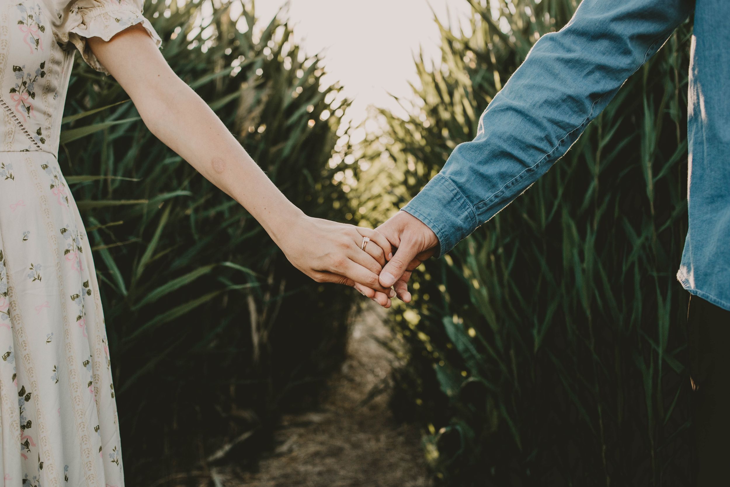Two people in blue and white clothing hold hands while surrounded by tall green plants in a garden setting.