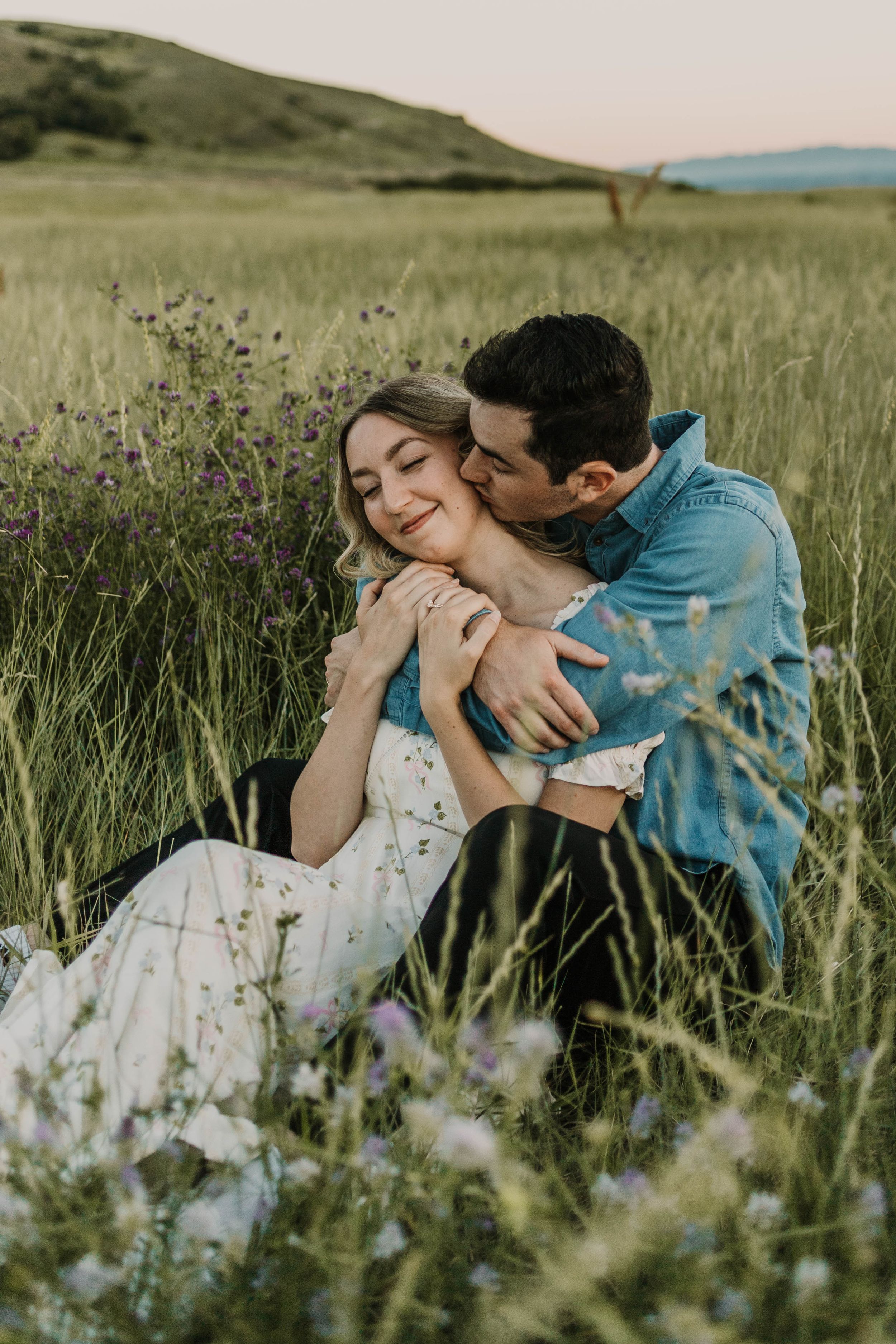 A romantic couple embraces in a sunlit meadow with wildflowers and tall grass during a summer photoshoot.