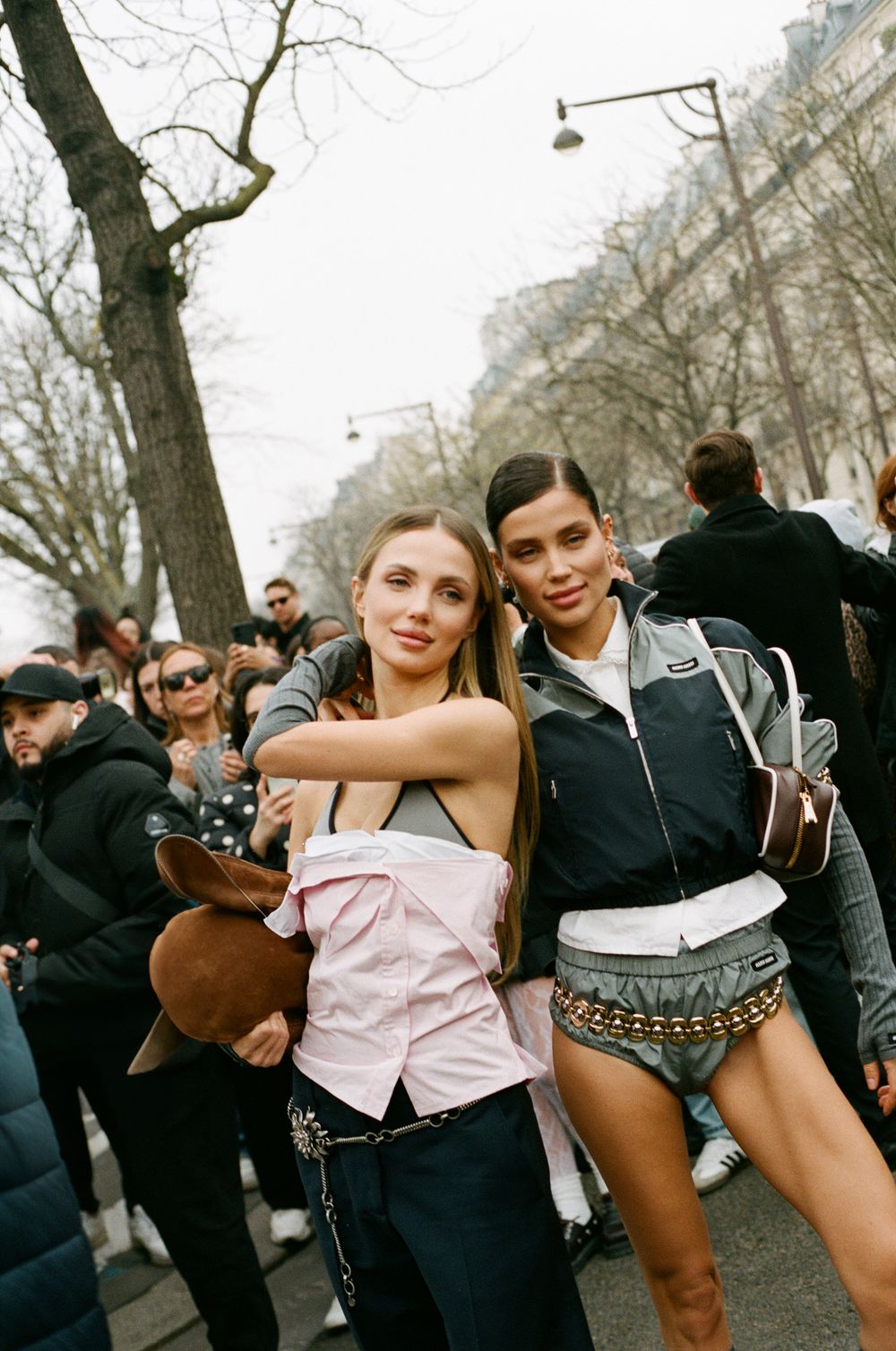 Two young people pose together with casual style at an outdoor gathering on a tree-lined street during fashion week.