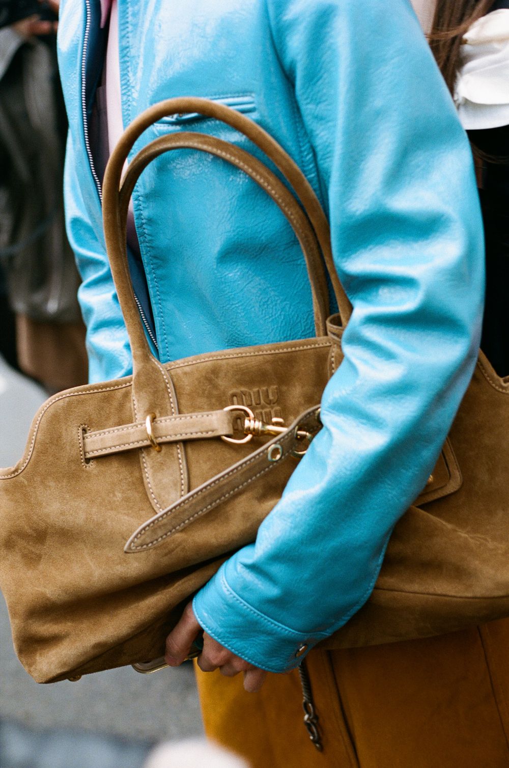 Person wearing bright turquoise sweater carries a tan suede handbag with leather handles against urban background.