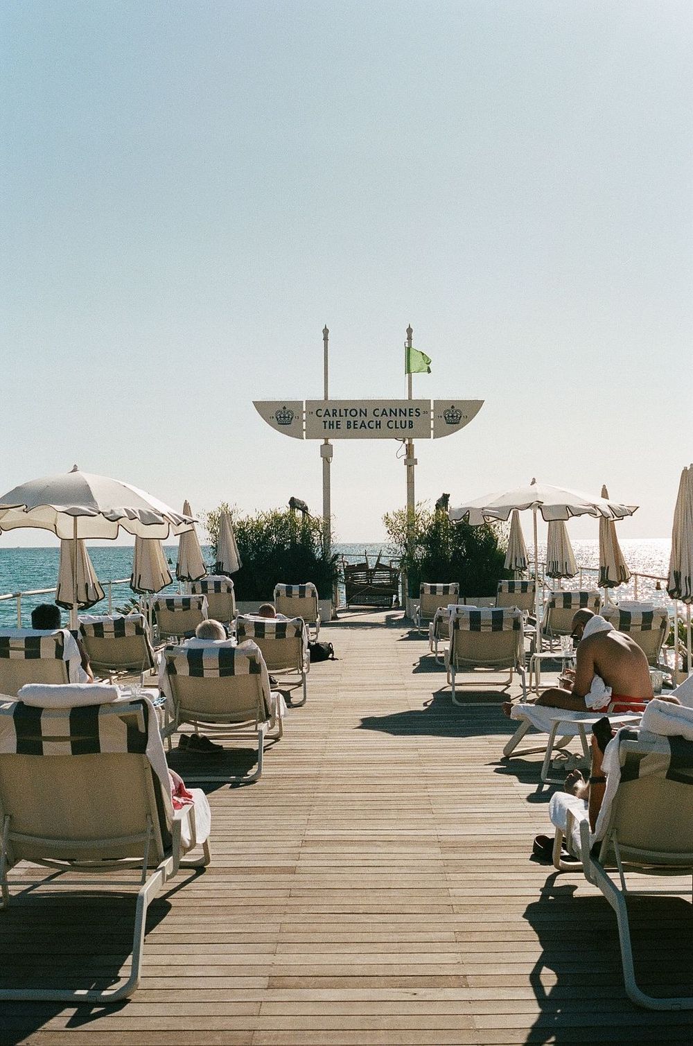 Beach lounge chairs line a wooden boardwalk with umbrellas and ocean views at a seaside resort during daytime.