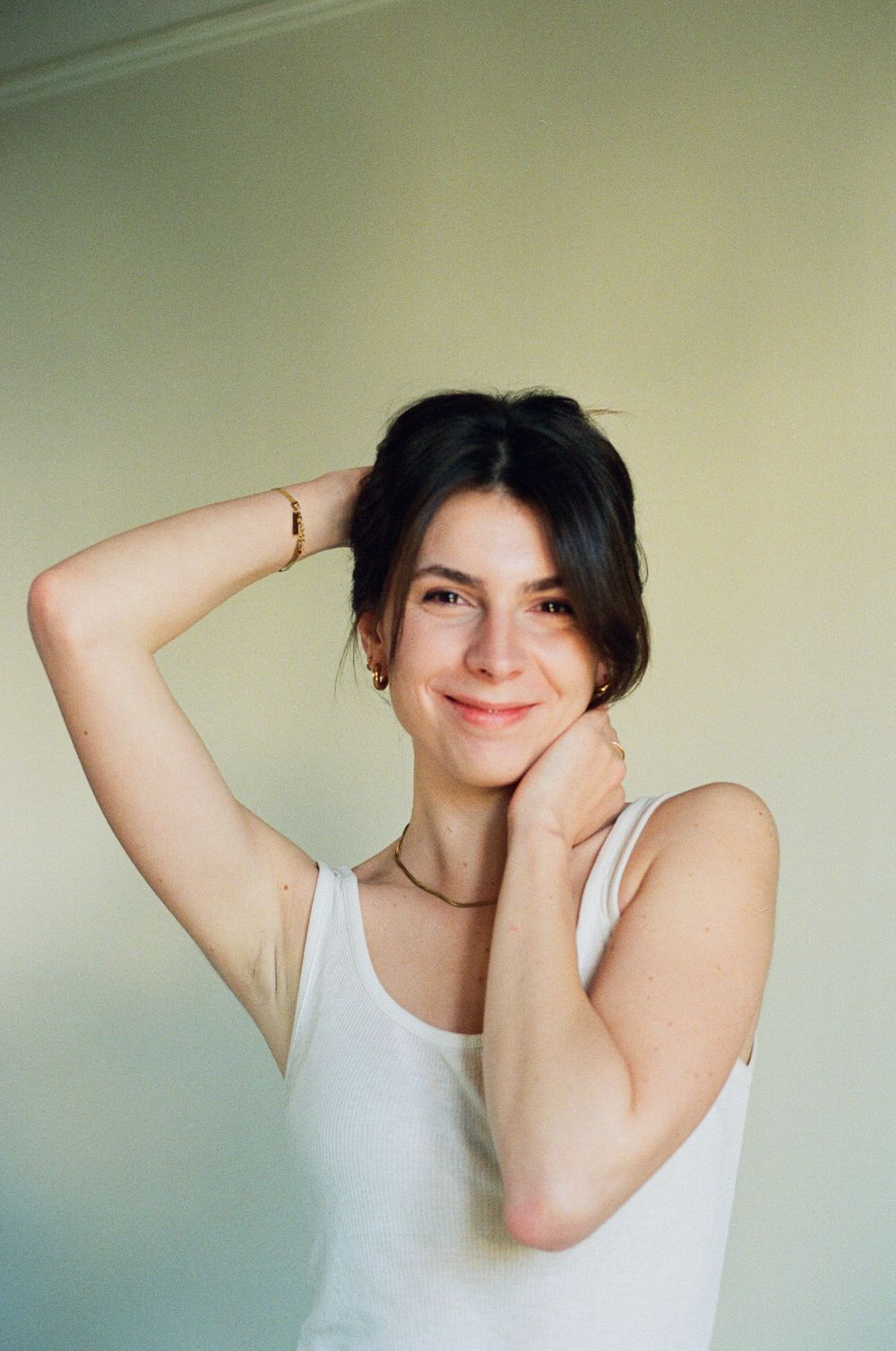 Portrait of person in white tank top smiling with hands in relaxed pose against light background.