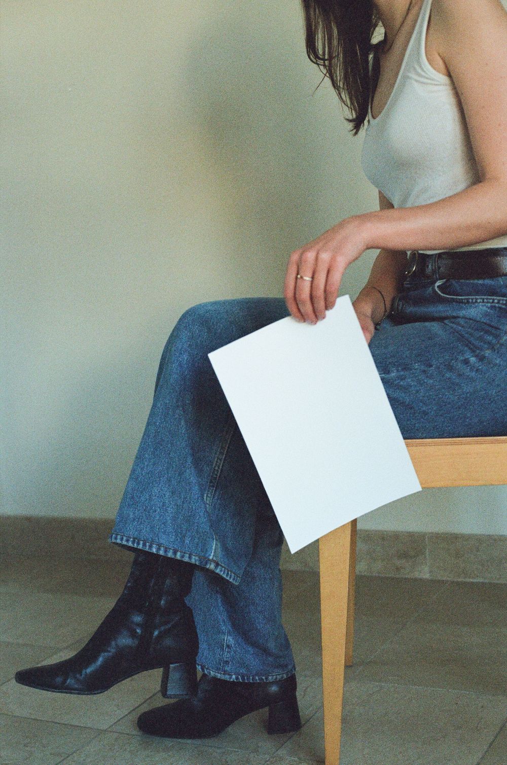 Person in white tank top and jeans sits holding a blank white paper while wearing black boots.