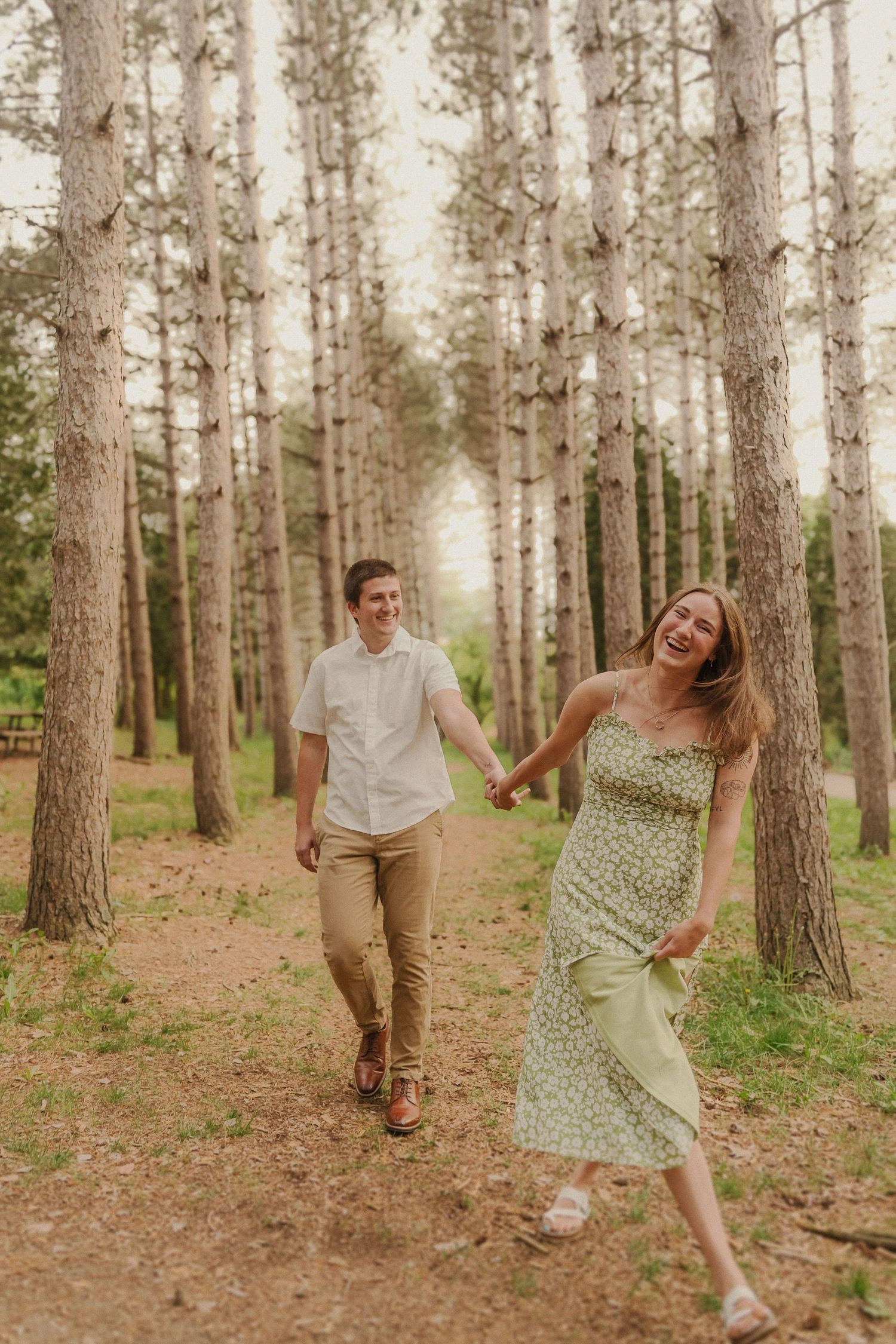 Couple playfully walking hand-in-hand through tall pine trees on a forest trail.