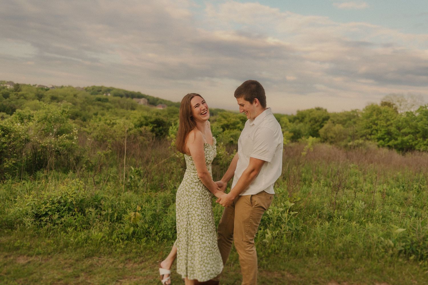 A couple stands together in a scenic meadow at sunset wearing casual summer attire against a backdrop of rolling hills.