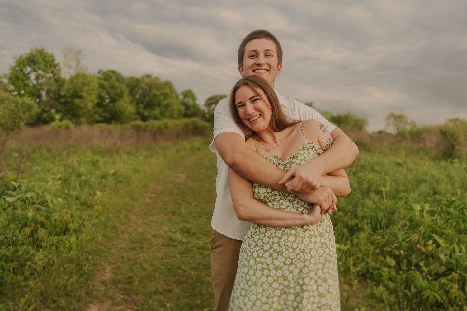 A couple embraces in a loving moment on a grassy path during golden hour with green trees in the background.