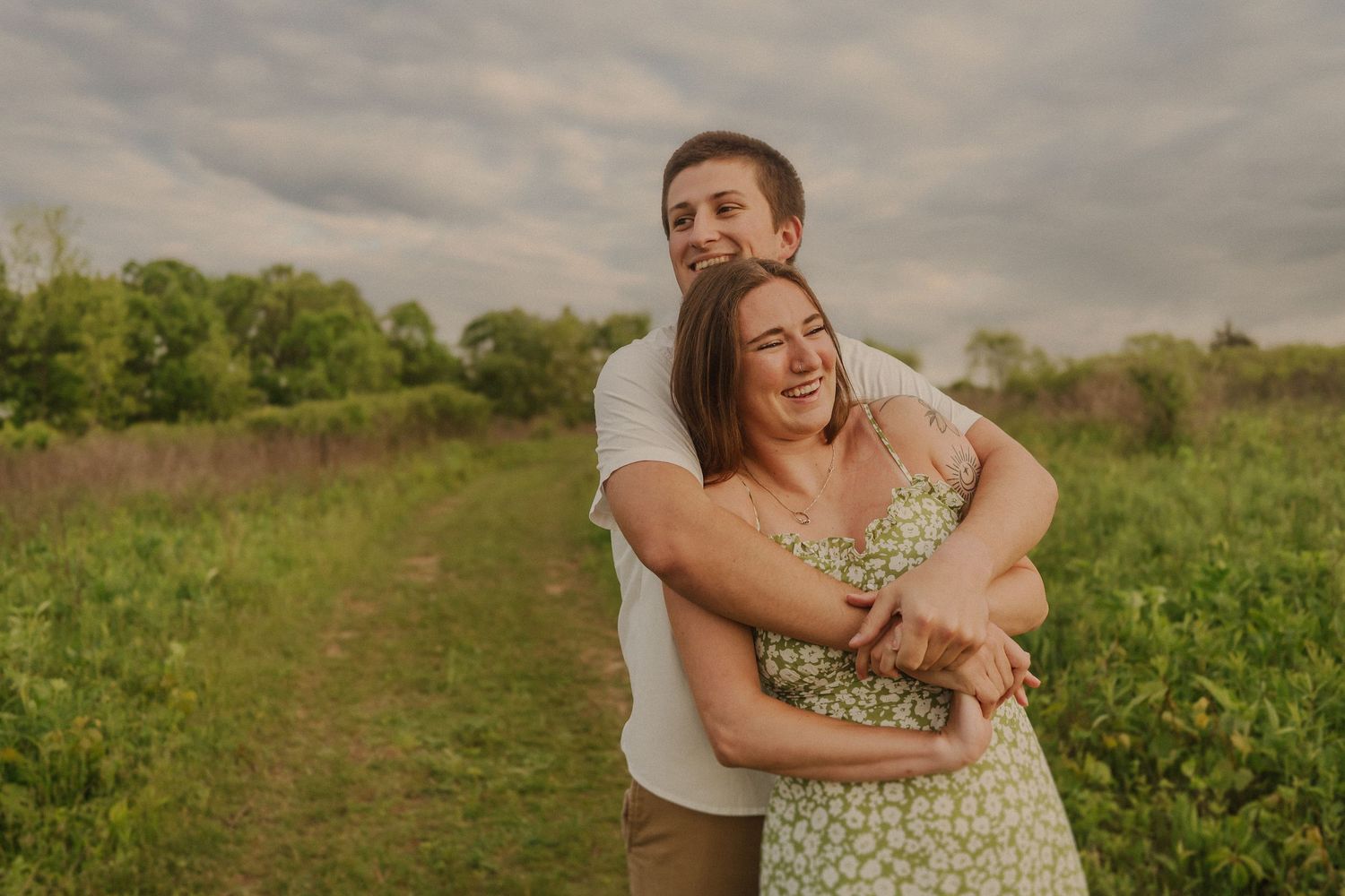 A couple embraces and laughs together in a sunny meadow with wildflowers and green trees in the background.