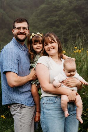 A family of four poses together outdoors in a meadow with wildflowers and evergreen trees in the background.