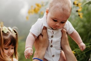 A person holds and interacts with a smiling baby outdoors in a natural setting with sunlit foliage in the background.
