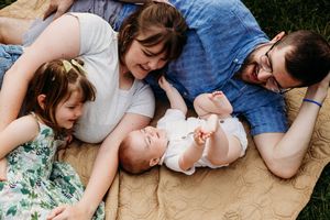 A family lies together on a tan blanket outdoors, smiling and looking down at a baby in a white outfit.