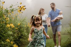 Young girl in floral dress runs joyfully through yellow wildflowers while family walks behind in misty summer meadow.
