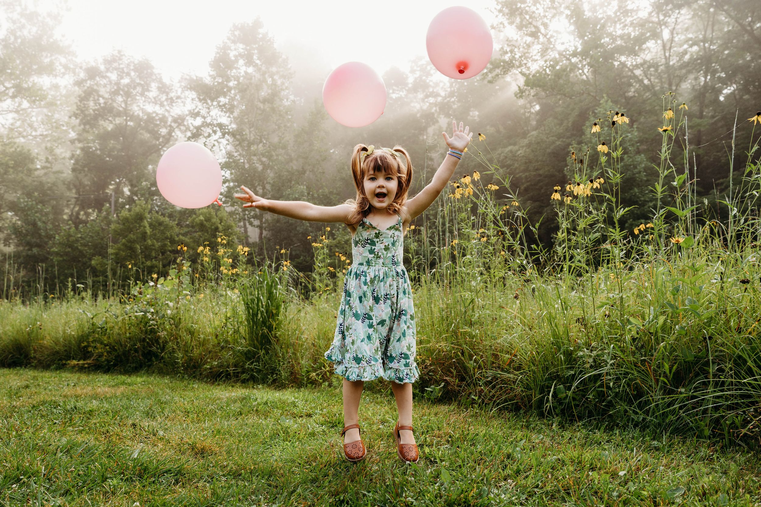 A child in a floral dress plays joyfully with pink balloons in a misty summer garden.