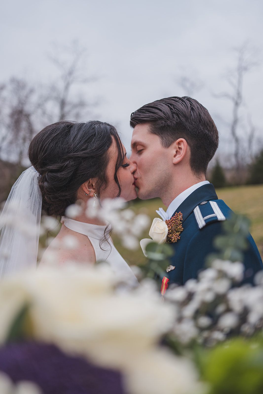 A tender wedding kiss shared between newlyweds, with the groom in military dress uniform and a romantic blurred floral foreground.