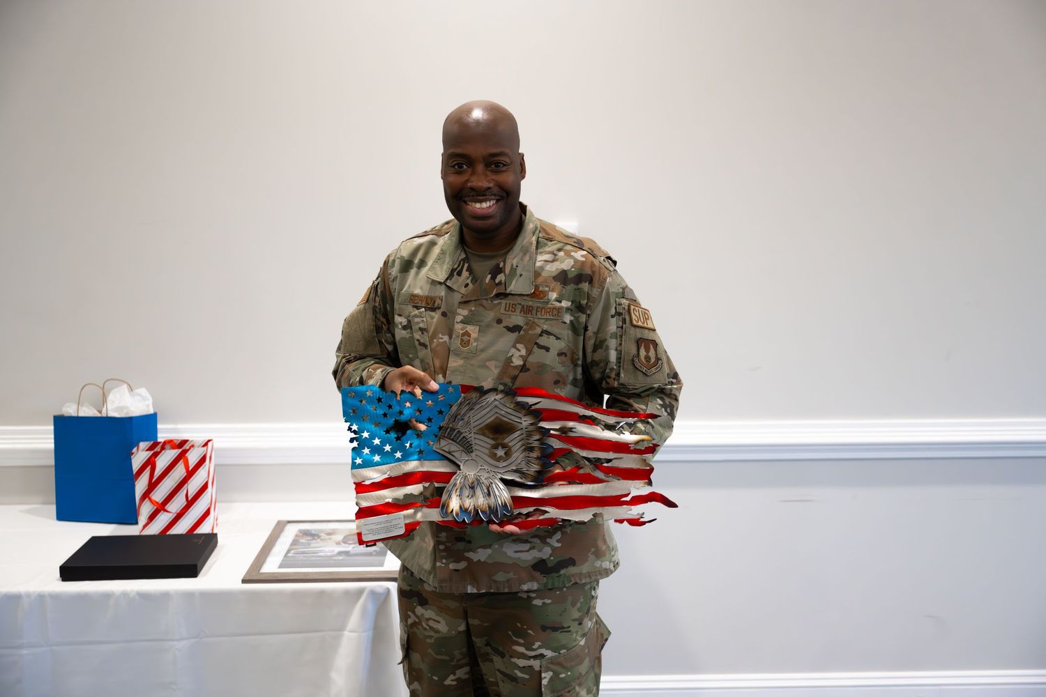 Military service member in camouflage uniform holds American flag art piece in front of white wall.