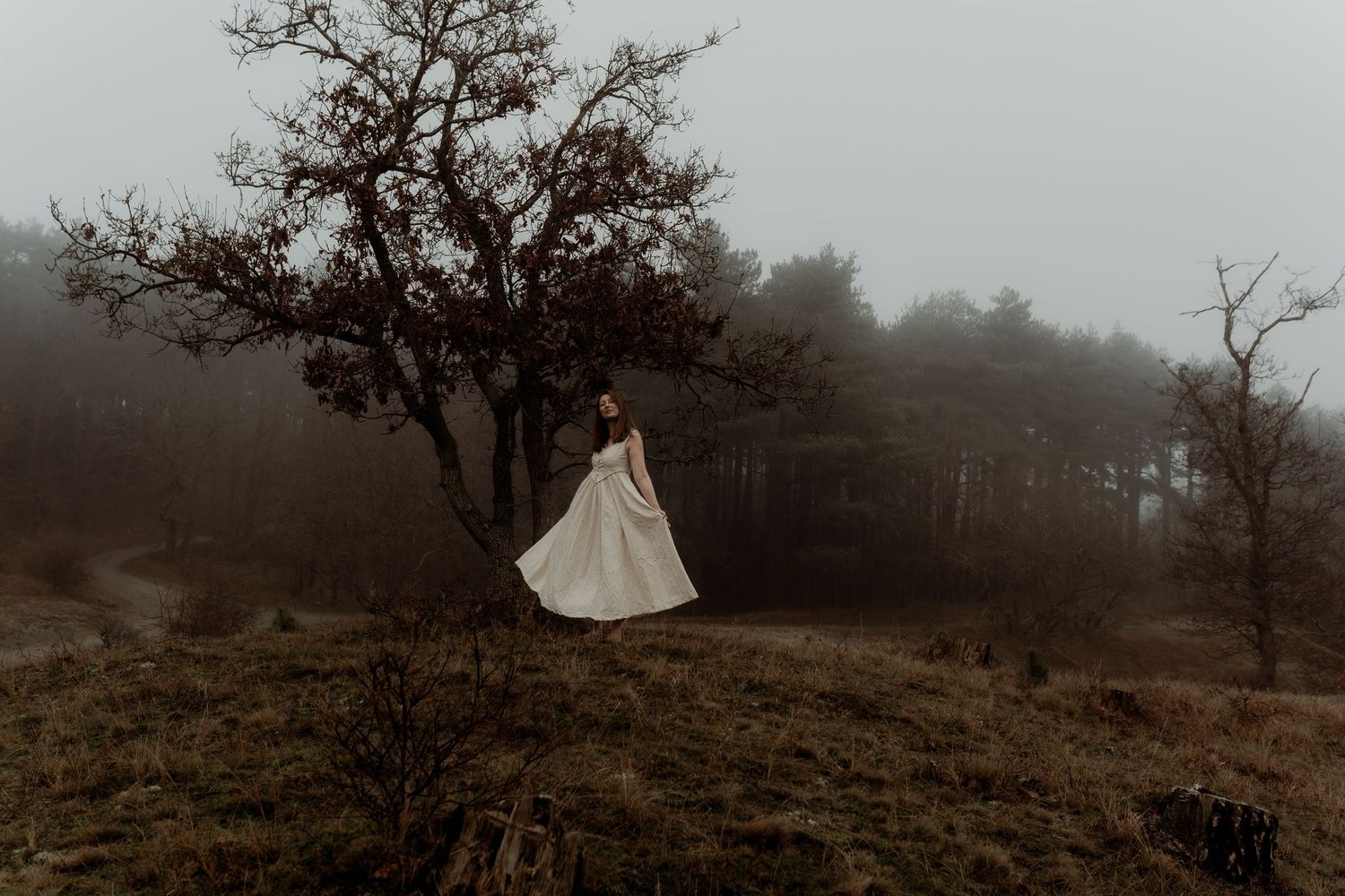 A person in a flowing emerald green dress stands on a misty Scottish moor with a white cottage in the background.