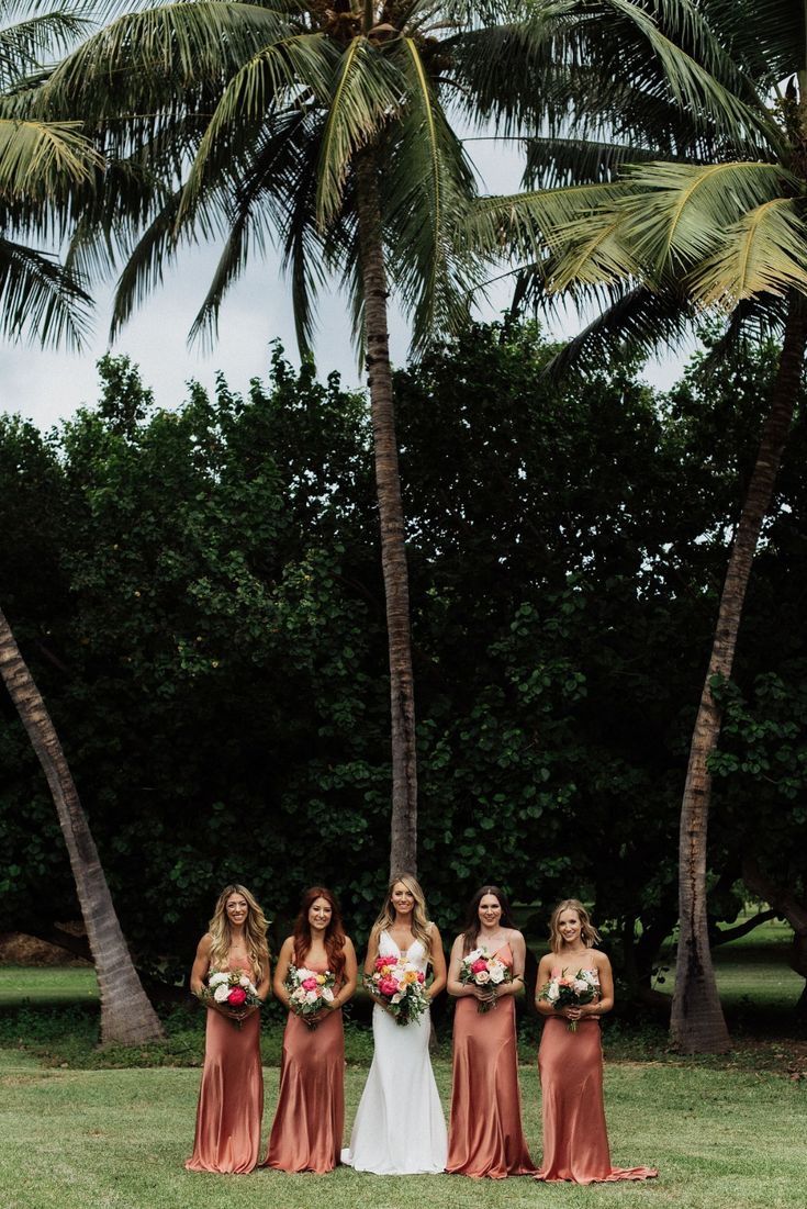 A group stands in a tropical setting with palm trees, wearing rose-colored gowns and holding bouquets on a grassy lawn.