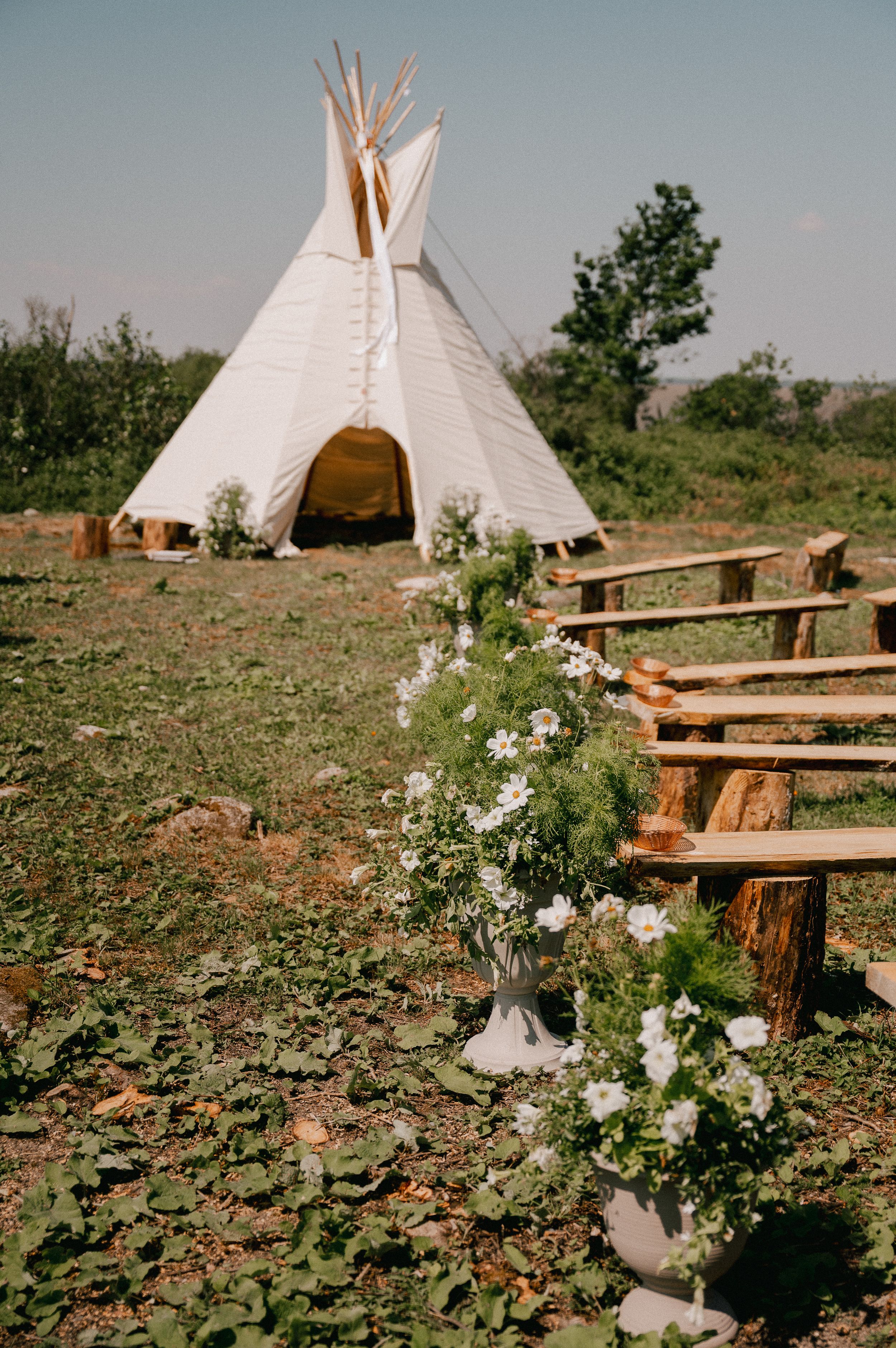 White teepee tents set up in a natural outdoor setting with rustic wooden benches and floral arrangements decorating the space.