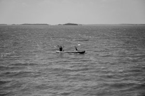 Two people paddle a small boat across choppy waters with distant islands visible on the horizon in black and white.