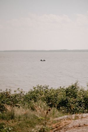 A small boat with two people floats on a serene lake or bay with distant shoreline visible through misty conditions.