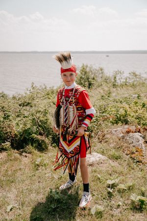 A young person wearing traditional Native American regalia plays by the coastline on a sunny day.