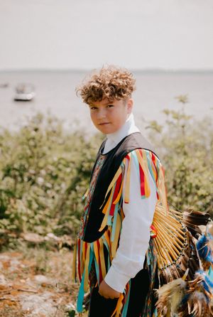 Colorful rainbow fringe adorns a young performer's traditional Native American regalia against a sunlit outdoor backdrop.