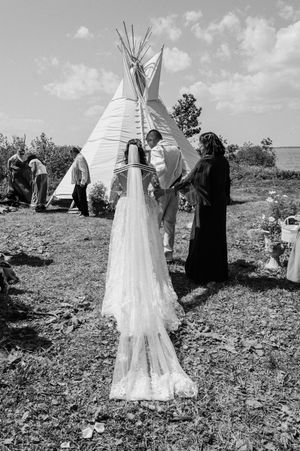 A black and white photograph of a rural wedding ceremony taking place near a traditional teepee structure in a field.