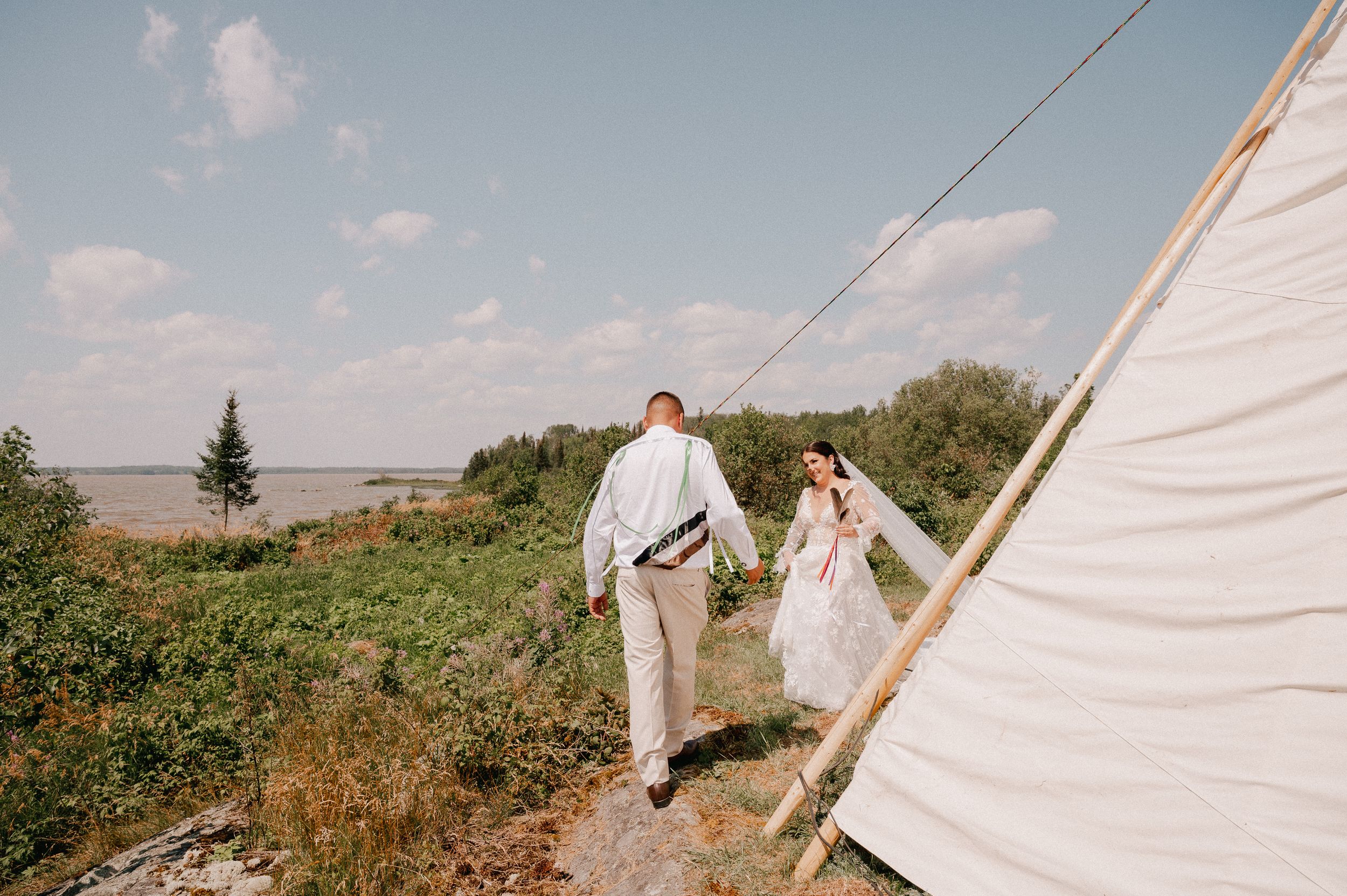 A couple walks together on grass near a white teepee structure under a bright blue sky.